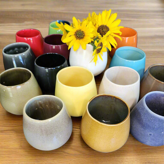 Ceramic wine cups in yellow, grey, blue, white, mustard, orange, black, wine, red, green, black and brown on wood table with yellow wildflowers in center white cup.