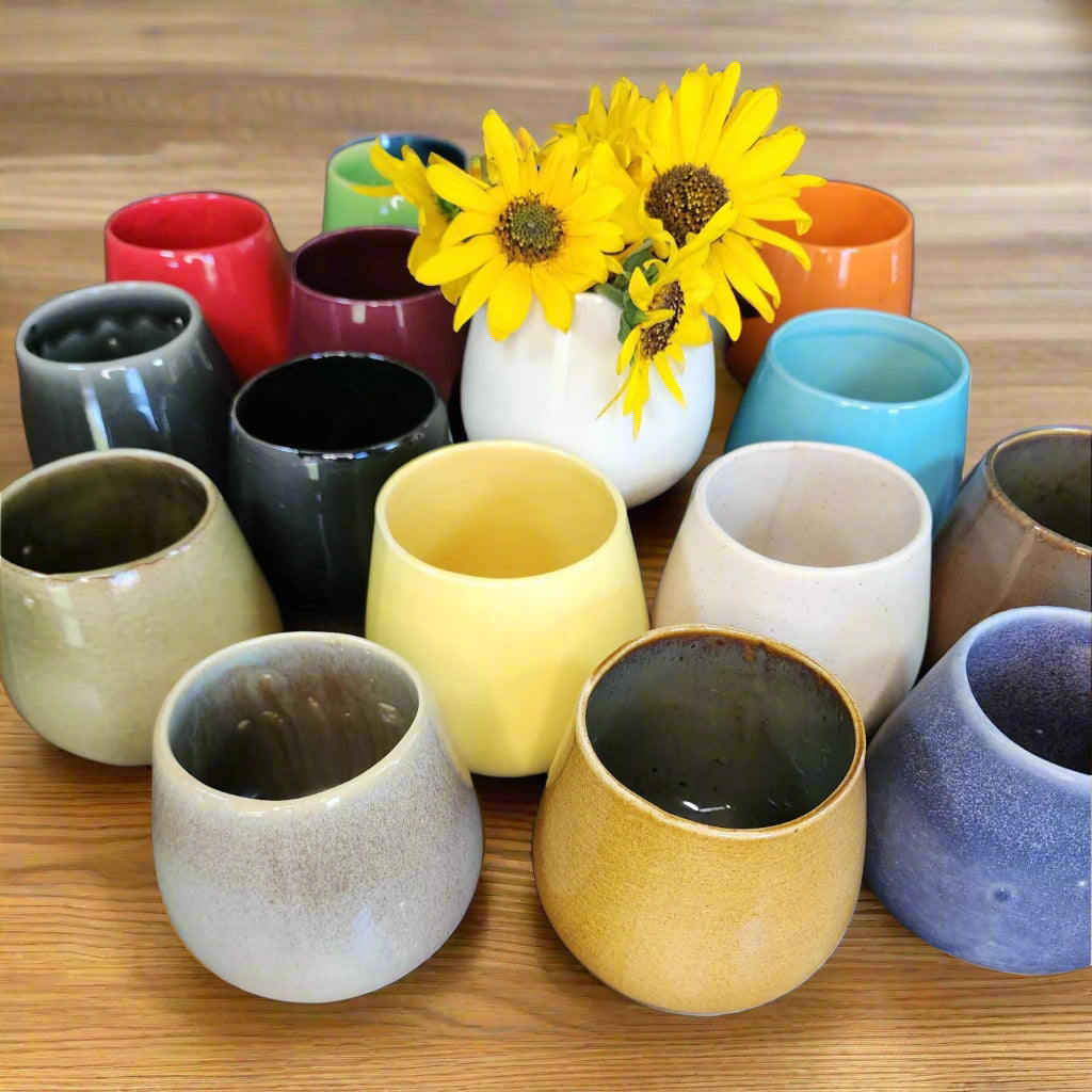 Ceramic wine cups in yellow, grey, blue, white, mustard, orange, black, wine, red, green, black and brown on wood table with yellow wildflowers in center white cup.
